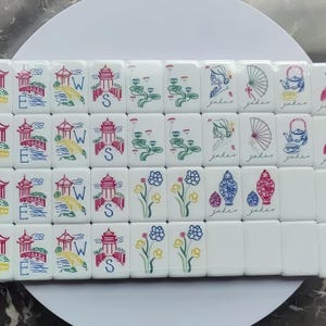 May include: A collection of white Mahjong tiles with colorful illustrations. The tiles feature images of pagodas, bridges, flowers, and teapots. Some tiles have the letter "S" or the word "joker" on them.