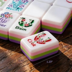 May include: Close-up of colourful Mahjong tiles. The tiles are white with a layered design of pink, green, and purple. The tiles have various illustrations and text, including "Crak" and "Bam". The tiles measure 3.1 cm x 2.3 cm x 1.5 cm.