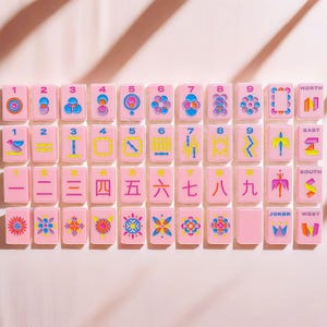May include: A set of rectangular Mahjong tiles in a soft pink color, featuring colorful engraved designs including numbers one through nine, directional markers, floral patterns, and a Joker tile with a crown graphic, all arranged in a grid.