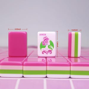 May include: A close-up of pink and white Mahjong tiles. One tile is solid pink, measuring 0.95 inches wide. Another tile features a woman graphic and the word "JOKER". A third tile has pink, green, and white stripes, measuring 0.63 inches wide. The tiles are 1.26 inches tall.