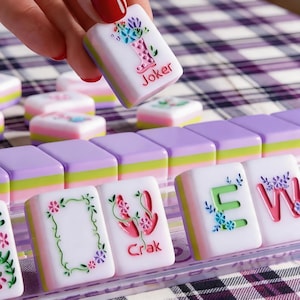 May include: A set of colourful Mahjong tiles. One tile is held up, displaying a floral design and the word "Joker." Other tiles show various symbols and letters. The tiles are white with pastel accents and are arranged on a plaid surface.
