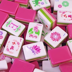May include: A collection of pink and white Mahjong tiles. The tiles feature various symbols and characters in pink, green, and white. Some tiles have pink and green stripes on the sides. The tiles are scattered, creating a visually appealing arrangement.