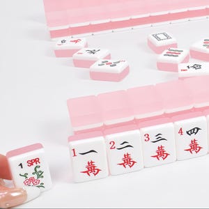 May include: A set of light pink and white Mahjong tiles featuring black and red characters, numbers, and floral designs. The tiles are arranged in rows and scattered, with one tile held up showing the text 1 SPR and a green floral illustration.