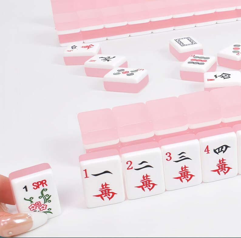 May include: A set of light pink and white Mahjong tiles featuring black and red characters, numbers, and floral designs. The tiles are arranged in rows and scattered, with one tile held up showing the text 1 SPR and a green floral illustration.