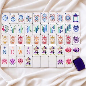 May include: A full Mahjong set with white tiles displaying colourful symbols, numbers, and characters. The set includes tiles with floral designs, bamboo, and the word "NEWS". A purple tile holder is also included.