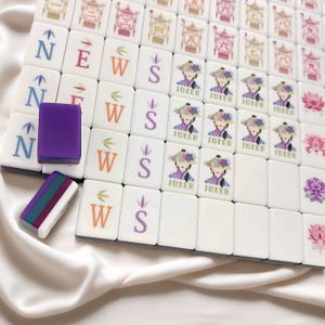 May include: A collection of white Mahjong tiles, displaying a variety of symbols, characters, and illustrations. The tiles show the letters N, E, W, and S, alongside images of a woman, flowers, and Chinese characters. Two purple and multi-coloured tile holders are also visible.