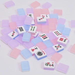 May include: A collection of rectangular mahjong tiles in translucent pastel shades of pink, blue, and purple. Some tiles feature printed numbers, symbols, and characters in black, red, and green ink, including a Joker tile and a floral design.