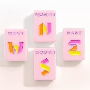 May include: Four rectangular light pink tiles arranged in a compass formation. Each tile features a cardinal direction label in purple text and a stylized letter icon in orange, yellow, and magenta. The tiles are labeled NORTH, SOUTH, EAST, and WEST.