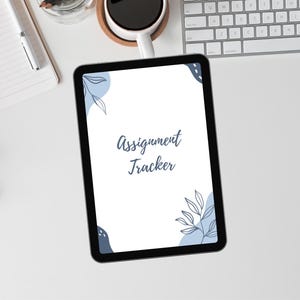 Peut inclure: Une tablette noire affichant le texte "Assignment Tracker" dans une police manuscrite. L'écran de la tablette présente des motifs floraux bleus. La tablette est posée sur une surface blanche, avec un carnet, un stylo et une tasse de café visibles.