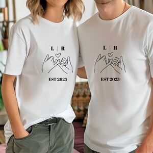 Könnte beinhalten: Weiße T-Shirts mit einer schwarzen Strichzeichnung von zwei Händen, die sich beim Schwur mit dem kleinen Finger verschränken, einem Herz und dem Text "L R" darüber und "EST 2023" darunter. Die Shirts werden von zwei Personen getragen.