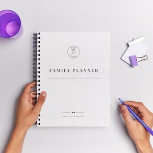 May include: A white spiral-bound Family Planner with the text "Family Planner" and "Organize Your Family Life With Ease". A purple pen and a purple cup are also visible.