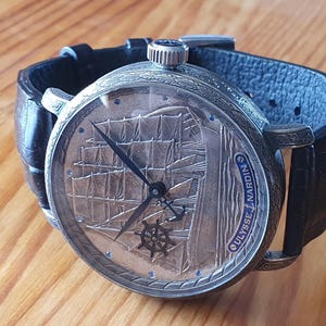 Może przedstawiać: Srebrny zegarek z czarnym sk&oacute;rzanym paskiem. Tarcza zegarka przedstawia szczeg&oacute;łowy grawerunek żaglowca oraz napis "ULYSSE NARDIN". Zegarek leży na drewnianej powierzchni.
