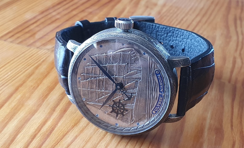 Może przedstawiać: Srebrny zegarek z czarnym sk&oacute;rzanym paskiem. Tarcza zegarka przedstawia szczeg&oacute;łowy grawerunek żaglowca oraz napis "ULYSSE NARDIN". Zegarek leży na drewnianej powierzchni.