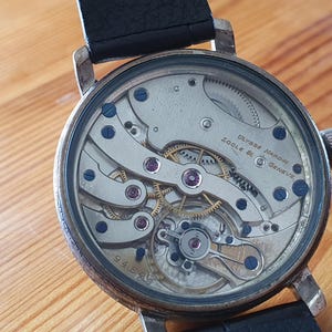 Może przedstawiać: Zbliżenie zabytkowego zegarka Ulysse Nardin z czarnym sk&oacute;rzanym paskiem. Tarcza zegarka odsłania skomplikowane elementy mechaniczne, w tym koła zębate, śruby i klejnoty. Widoczny jest napis "Ulysse Nardin Locle & Geneve".