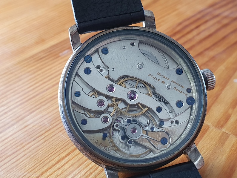 Może przedstawiać: Zbliżenie zabytkowego zegarka Ulysse Nardin z czarnym sk&oacute;rzanym paskiem. Tarcza zegarka odsłania skomplikowane elementy mechaniczne, w tym koła zębate, śruby i klejnoty. Widoczny jest napis "Ulysse Nardin Locle & Geneve".