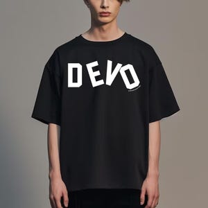 May include: Black t-shirt with the word "DEVO" in large, white, block letters across the chest. The shirt has short sleeves and a crew neck. The text is slightly angled. The shirt is worn by a person.