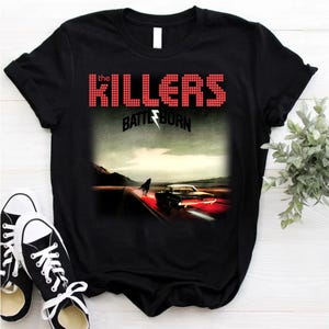 May include: Black t-shirt featuring the band name "The Killers" in red pixelated font above the album title "Battle Born". The graphic depicts a car on a road with a horse silhouette. Black and white sneakers are in the foreground.
