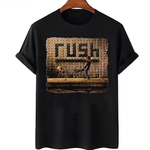 May include: Black t-shirt with a graphic of a person kicking a football, with the word "RUSH" in large letters. The graphic has a vintage, textured appearance. The shirt is made of a soft material and has short sleeves.