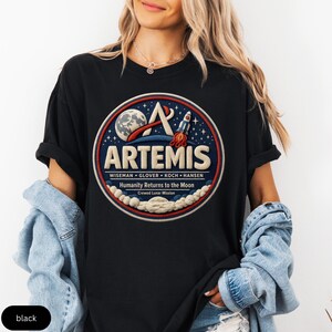 May include: Black crew neck t-shirt with a circular Artemis design. The design includes a rocket, moon, and stars, along with the text "ARTEMIS" and "Humanity Returns to the Moon." The shirt is a casual style.