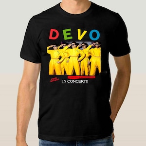 May include: Black t-shirt featuring the band name "DEVO" in colorful letters. The shirt displays a graphic of the band members in yellow jumpsuits, saluting. Text below reads "FOUR SHOWS!! IN CONCERT!!"