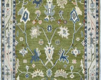 May include: A rectangular area rug featuring a vibrant olive green field adorned with intricate, symmetrical floral and botanical motifs in shades of blue, cream, and soft red. The rug is framed by a multi-layered border with geometric patterns.