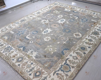 May include: A large rectangular area rug featuring a grey field with a repeating pattern of floral and geometric motifs in shades of blue, cream, and tan. The rug has a wide cream-coloured border with intricate floral and vine designs.