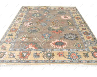 May include: A rectangular area rug featuring a taupe central field adorned with a floral pattern in shades of coral, blue, and green. The rug is framed by a wide beige border with a repeating floral motif and finished with white fringe.