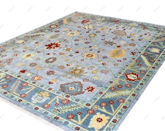 May include: A rectangular area rug featuring a light blue field decorated with a repeating pattern of floral and botanical motifs in shades of red, yellow, green, and brown, framed by a wide border with similar floral designs and fringe.