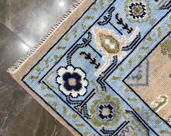 May include: A rectangular area rug featuring a light blue border with a repeating floral pattern in dark blue, green, and cream. The central field is a solid beige colour. The rug has a short, cream-coloured fringe along the edge.