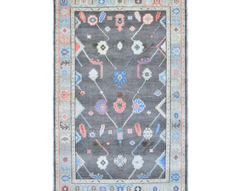 May include: A rectangular area rug with a dark grey field featuring a symmetrical pattern of stylised floral and geometric motifs in shades of light blue, coral pink, and white, enclosed by a decorative border with repeating patterns.