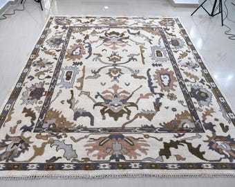 May include: A rectangular area rug featuring an off-white background with a symmetrical pattern of floral and geometric motifs in shades of brown, grey, and muted pink. The rug has a wide decorative border and a short fringe on the ends.