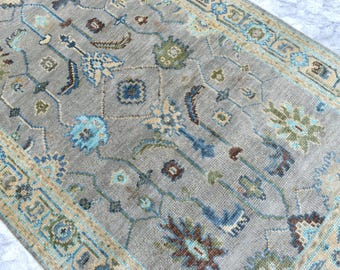 May include: A rectangular area rug featuring a grey field with a repeating geometric pattern in shades of blue, green, and tan. The rug has a wide border with intricate floral and geometric motifs in matching colours, finished with a textured pile.
