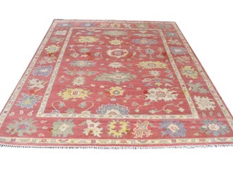 May include: A rectangular area rug featuring a deep red background with a repeating floral pattern in shades of blue, yellow, and cream. The rug has a wide decorative border and short white fringe along the top and bottom edges.