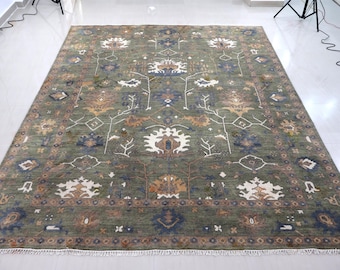 May include: A large rectangular area rug featuring an olive green background with a symmetrical floral pattern in shades of cream, tan, and navy blue. The rug has a wide decorative border and a short white fringe along the top and bottom edges.