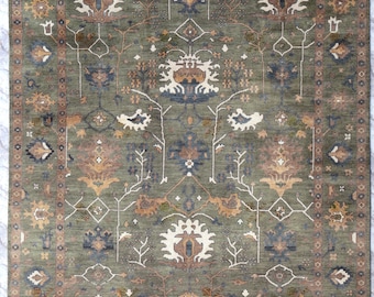 May include: A rectangular area rug featuring an olive green background with a symmetrical pattern of cream, tan, and navy blue floral and geometric motifs. The rug has a wide decorative border and a short white fringe along the edges.