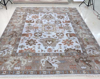 May include: A large rectangular area rug featuring a white central field with intricate geometric and floral motifs in shades of brown, tan, and muted orange. The rug has a wide, detailed border in matching earth tones and white fringe.