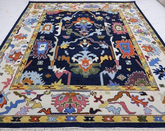 May include: A rectangular area rug featuring a deep navy blue central field with a symmetrical, multi-colored floral and geometric pattern in shades of orange, light blue, pink, yellow, and green, framed by a wide cream-colored border.
