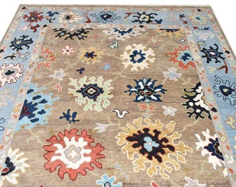 May include: A rectangular area rug featuring a taupe background with a repeating pattern of stylised floral motifs in shades of coral, yellow, navy blue, light blue, and white, framed by a light blue border with matching floral designs.