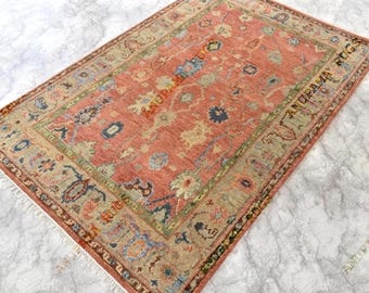 May include: A rectangular area rug featuring a coral-toned field with intricate floral and geometric patterns in shades of blue, green, and beige. The rug has a wide border and white fringe on the ends, with the text ANUPAMA RUGS visible.