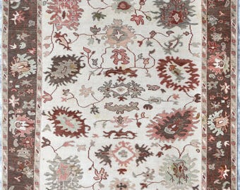 May include: A rectangular rug with a cream central field and a dark brown border. The rug features floral and geometric patterns in shades of brown, pink, and green. The border has a repeating floral design, and the rug has a fringed edge.