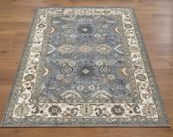 May include: A rectangular area rug featuring a central blue field with intricate floral and medallion patterns in shades of cream, beige, and dark grey. The rug is framed by a wide, ornate border with repeating botanical motifs.