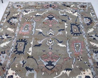 May include: A large rectangular area rug featuring a muted sage green background with a repeating pattern of stylised floral and geometric motifs in shades of navy blue, dusty pink, light blue, and cream, finished with a short fringe edge.
