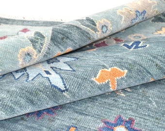 May include: A light blue area rug featuring a repeating pattern of stylised floral motifs in shades of orange, dark blue, and light purple. The rug has a thick, textured pile and is partially folded over to reveal the underside and weave.