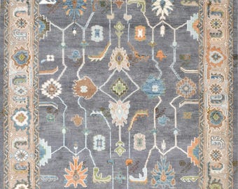 May include: A rectangular area rug with a dark grey field featuring a symmetrical pattern of stylised floral and geometric motifs in orange, blue, green, and cream. The rug has a wide border with repeating floral elements in similar colours.