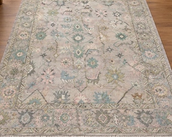 May include: A rectangular area rug featuring a light grey background with a repeating floral and geometric pattern in muted shades of sage green, dusty pink, and soft blue. The rug has a wide decorative border and short fringe edges.
