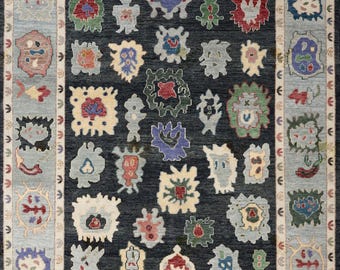 May include: A rectangular area rug featuring a dark charcoal central field filled with various abstract, multi-coloured shapes in red, green, blue, and cream. The rug is framed by a light blue border with repeating small motifs.
