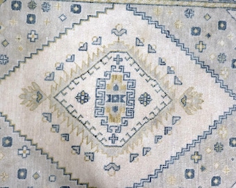 May include: A woven area rug featuring a light beige background with a large central diamond motif. The pattern includes geometric shapes, zig-zag borders, and small floral-like accents in shades of muted blue, soft gold, and cream.