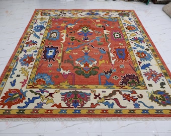 May include: A rectangular area rug featuring a vibrant terracotta red center field with intricate, multi-colored floral and geometric motifs in blue, green, yellow, and white. The rug has a wide cream-colored border with repeating patterns.