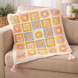 Könnte beinhalten: Eine gehäkelte Decke mit Patchwork-Design in Pastellfarben. Die Quadrate weisen gelbe, rosa und blaue Blumenmuster auf, die von Weiß umrandet sind. Die Decke hat einen weißen Rand und ist mit zwei Quasten in Rosa- und Gelbtönen verziert.