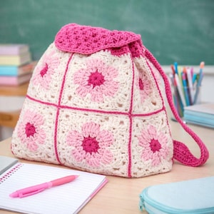 Op de afbeelding: Een handgemaakte gehaakte rugzak met een bloemenpatroon. De tas is gemaakt met crème, roze en fuchsia garen. De rugzak heeft een trekkoordsluiting en een schouderriem. De tas ligt op een bureau met een notitieboek en pen.